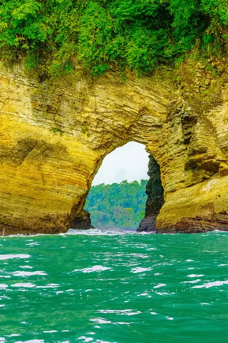 Rocky archway over turquoise water with lush greenery on top, leading to a view of distant trees.