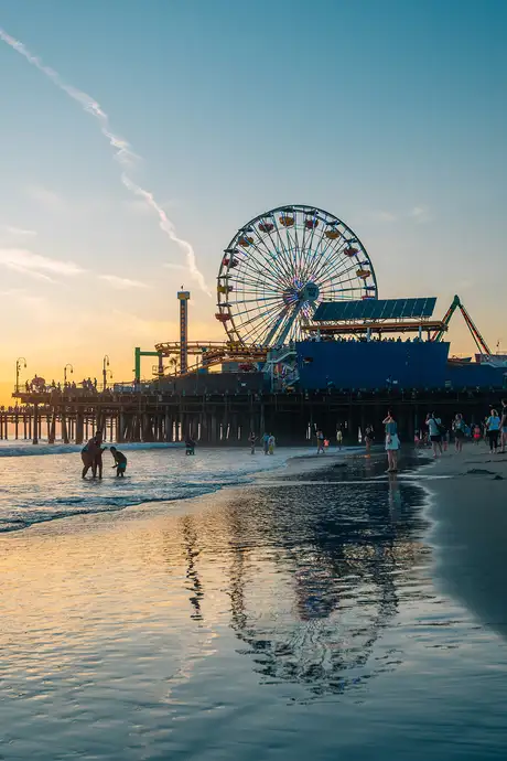 A beach at sunset with a Ferris wheel on a pier, people walking along the shore, and reflections in the wet sand.