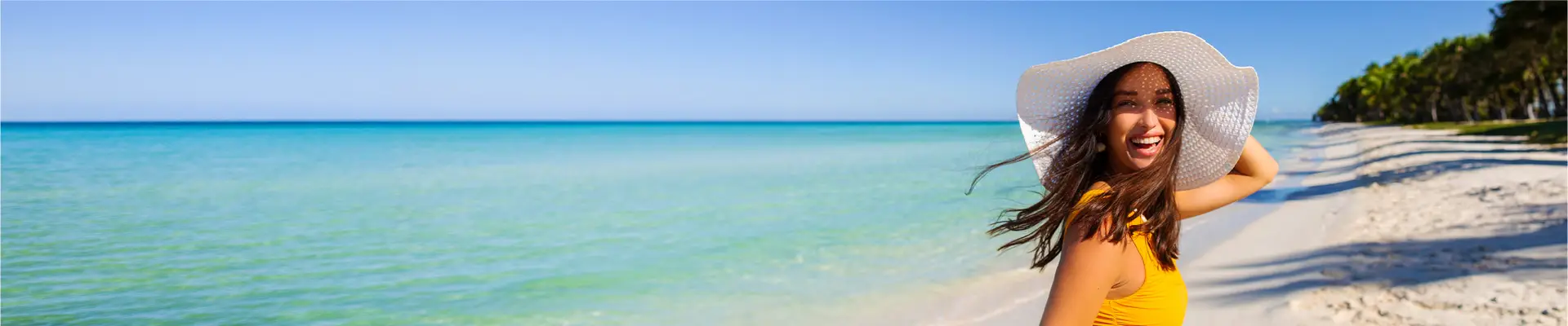A smiling woman in a yellow swimsuit and white sun hat stands on a tropical beach, with clear blue water and palm trees in the background.