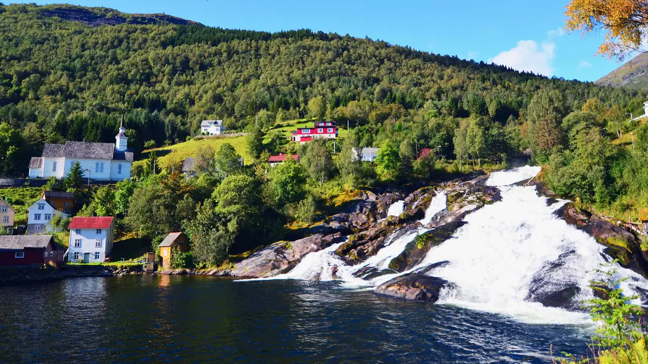 An image of Hellsylt in Geiranger, Norway. Showing a small number of houses in a wooded area next to a waterfall