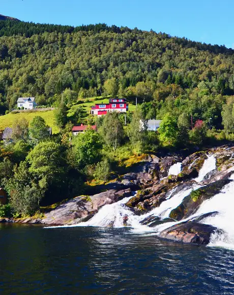 An image of Hellsylt in Geiranger, Norway. Showing a small number of houses in a wooded area next to a waterfall