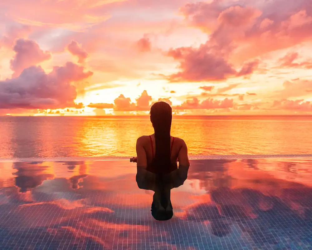 A woman in a pool looking out over the ocean at sunset