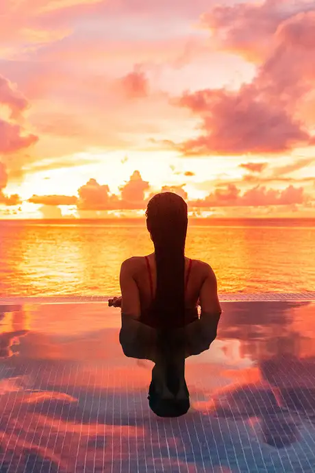 A woman in a pool looking out over the ocean at sunset