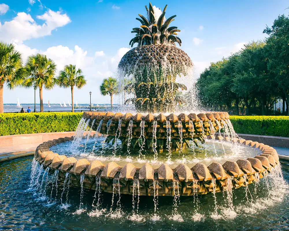 Pineapple-shaped fountain with cascading water, surrounded by palm trees and greenery, under a blue sky with scattered clouds.