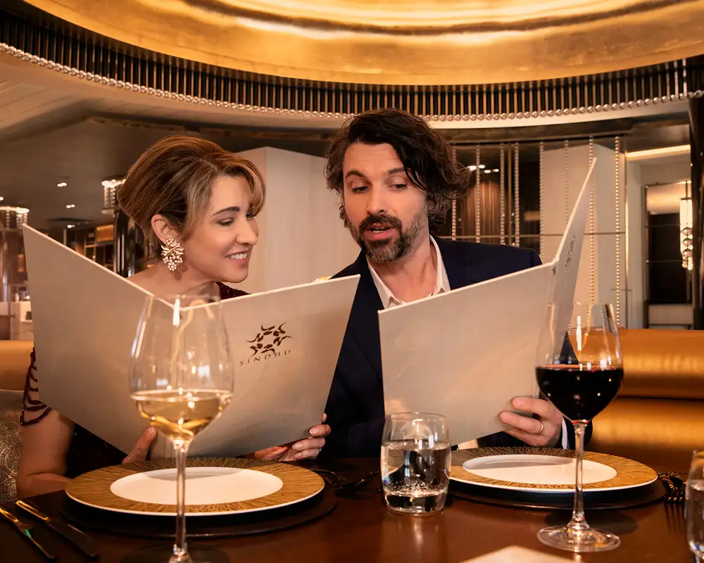 A couple sitting at a fancy restaurant table, looking at menus with wine glasses and tableware in front of them. Aboard a P&O Cruise Ship.