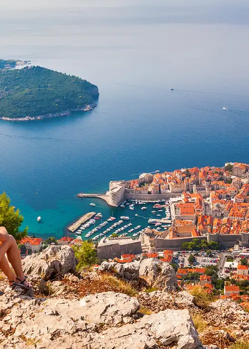 A woman sits on top of a cliff overlooking a sea side city in the Western Mediterranean