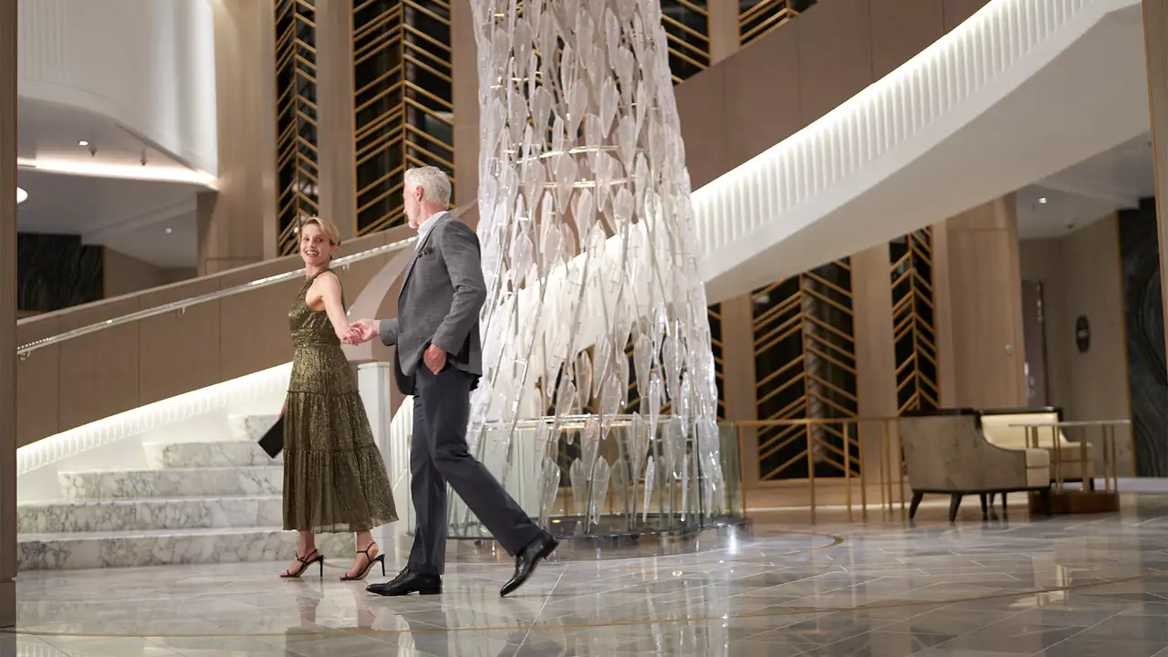 A couple elegantly dressed, walking hand in hand in a modern lobby with marble stairs and a tall decorative glass feature.