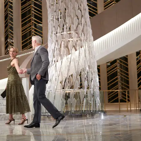 A couple elegantly dressed, walking hand in hand in a modern lobby with marble stairs and a tall decorative glass feature.