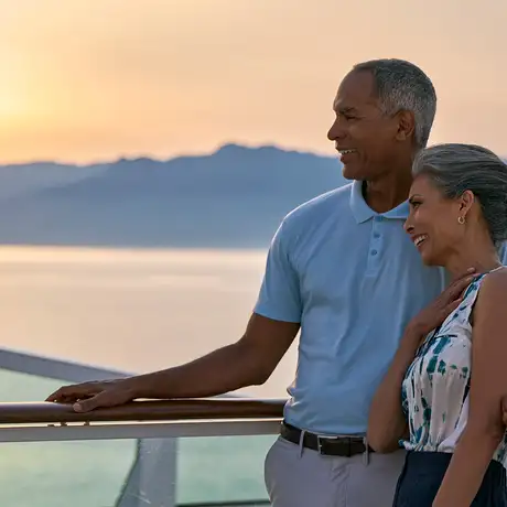 A smiling couple embraces on a ship's deck at sunset, with mountains and calm water in the background.