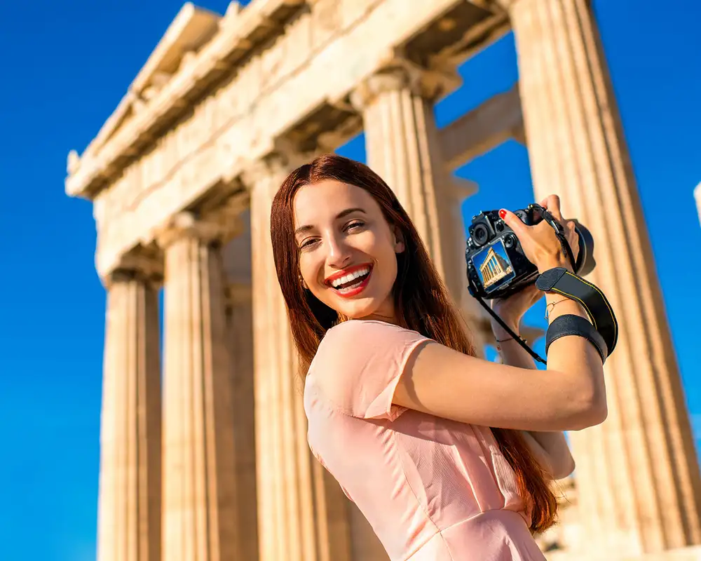 A Woman Taking a picture of the Acropolis in Athens, looking back at the camera smiling
