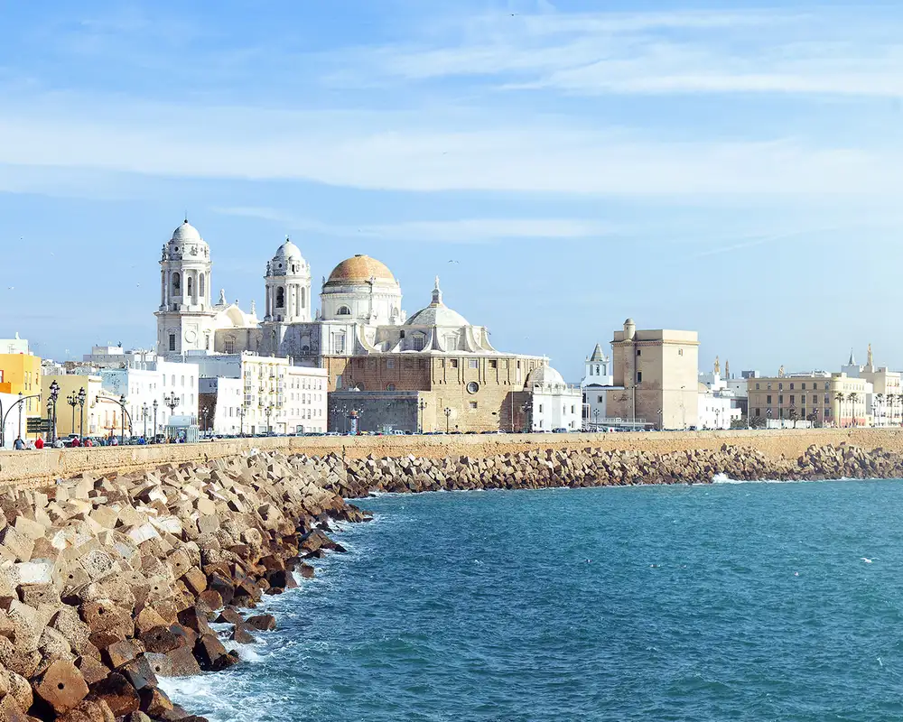 Coastal view of Cádiz, Spain, featuring the Cathedral with its distinctive domes, a stone seawall, and the blue waters of the Atlantic Ocean.