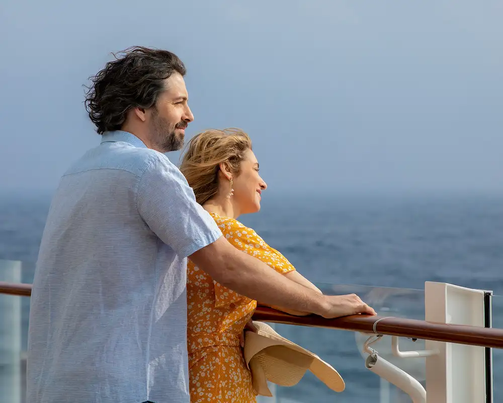 A couple stands on a ship's deck, enjoying the ocean view. The man wears a light blue shirt, and the woman holds a sunhat, wearing a yellow dress. Aboard a P&O Cruise Ship.