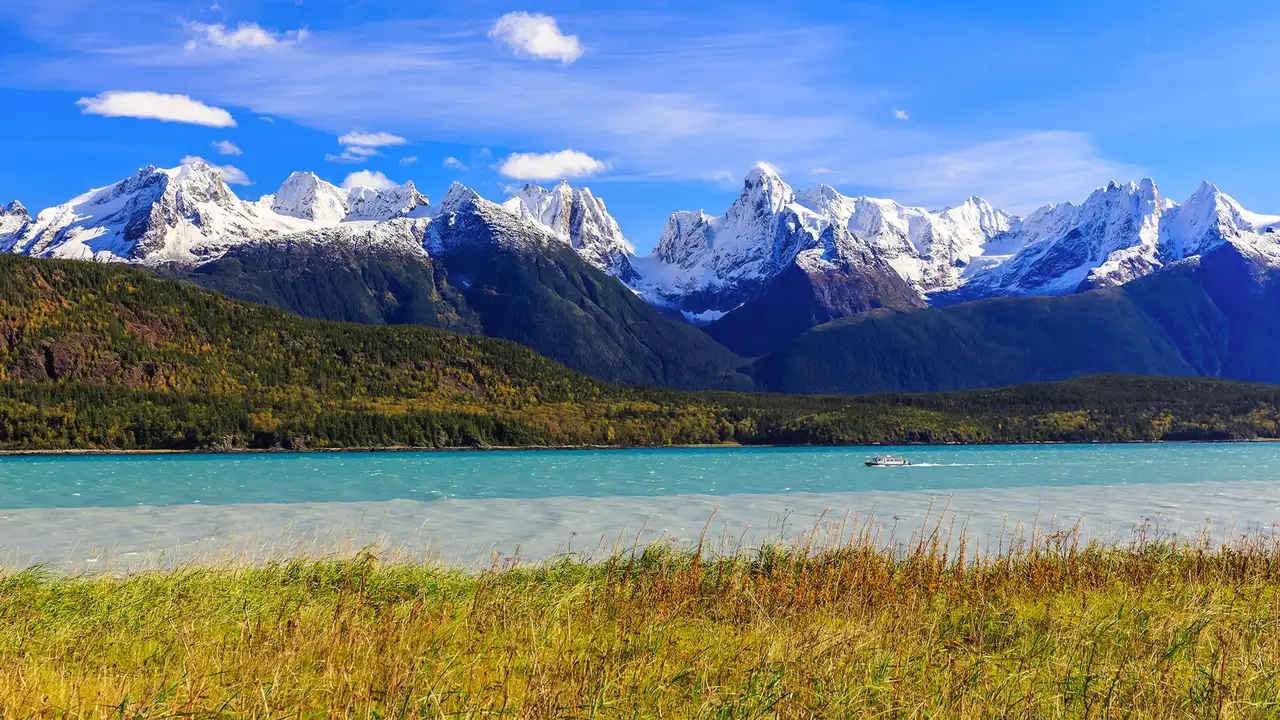 A shot of the landscape of Skagway, Alaska. With mountainrs visible in the background, a river in the middle and a grassy bank in the foreground