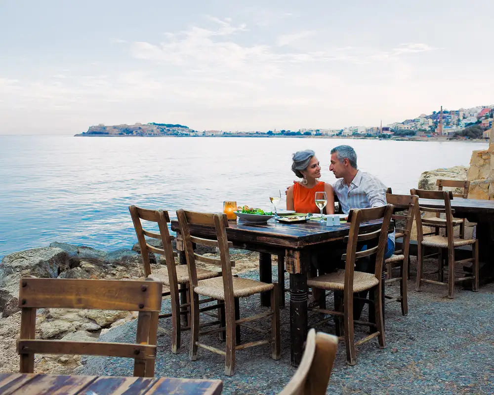 Couple dining at a seaside restaurant with a scenic view of the ocean and distant town, wooden tables and chairs surrounding them.