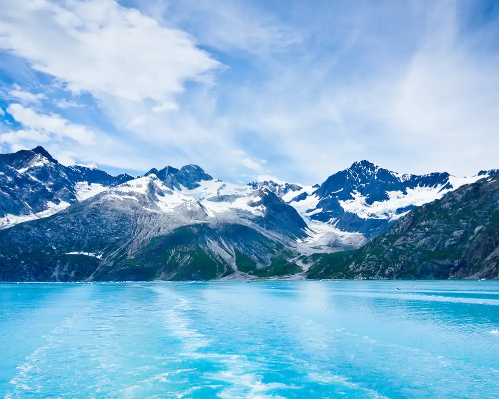 A stunning view of turquoise glacial waters with a backdrop of snow-capped mountains under a partly cloudy sky.