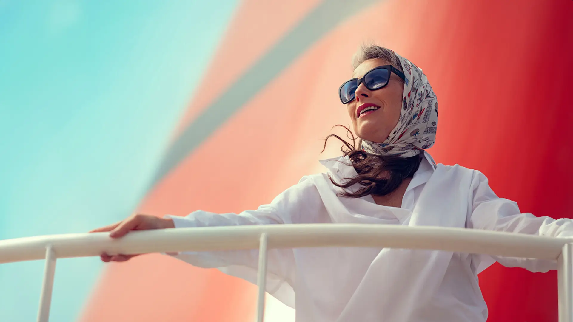 Woman wearing sunglasses and a scarf smiles, leaning on a white railing with a colorful background.