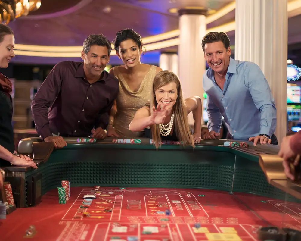 A group of people dressed in smart casual attire are gathered around a casino table, eagerly watching a game. A woman in pearls is reaching out animatedly. Aboard a Celebrity Cruise Ship