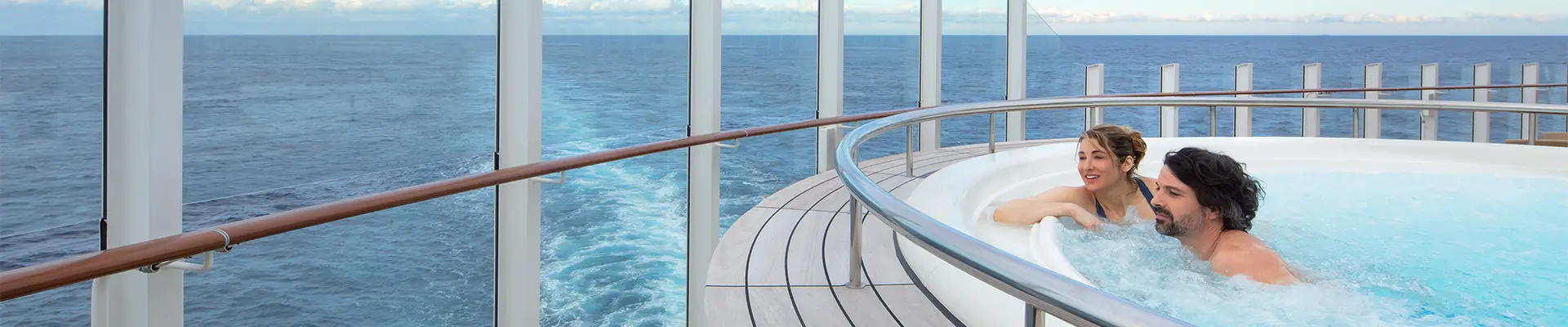 A couple enjoying a jacuzzi on a cruise ship deck, overlooking the blue ocean and clear sky, enclosed by glass panels. Aboard a P&O Cruise Ship.