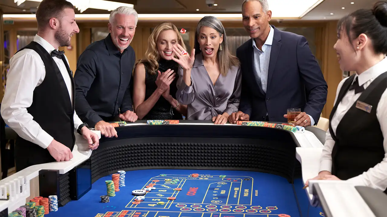 People excitedly playing craps at a casino table, with a woman in a gray blouse throwing dice, surrounded by smiling onlookers and dealers.