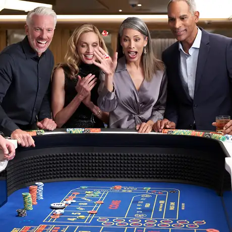 People excitedly playing craps at a casino table, with a woman in a gray blouse throwing dice, surrounded by smiling onlookers and dealers.
