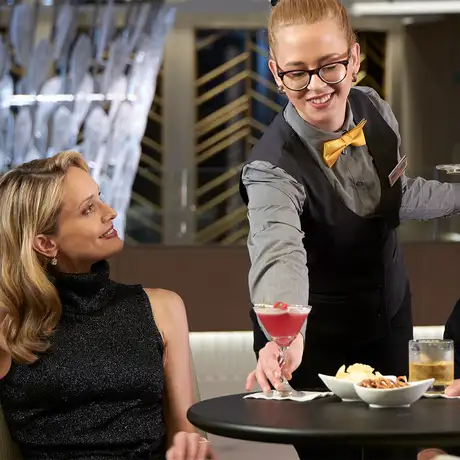 A waiter serves a cocktail to a smiling man and woman sitting at a table, with a stylish gold and glass background.