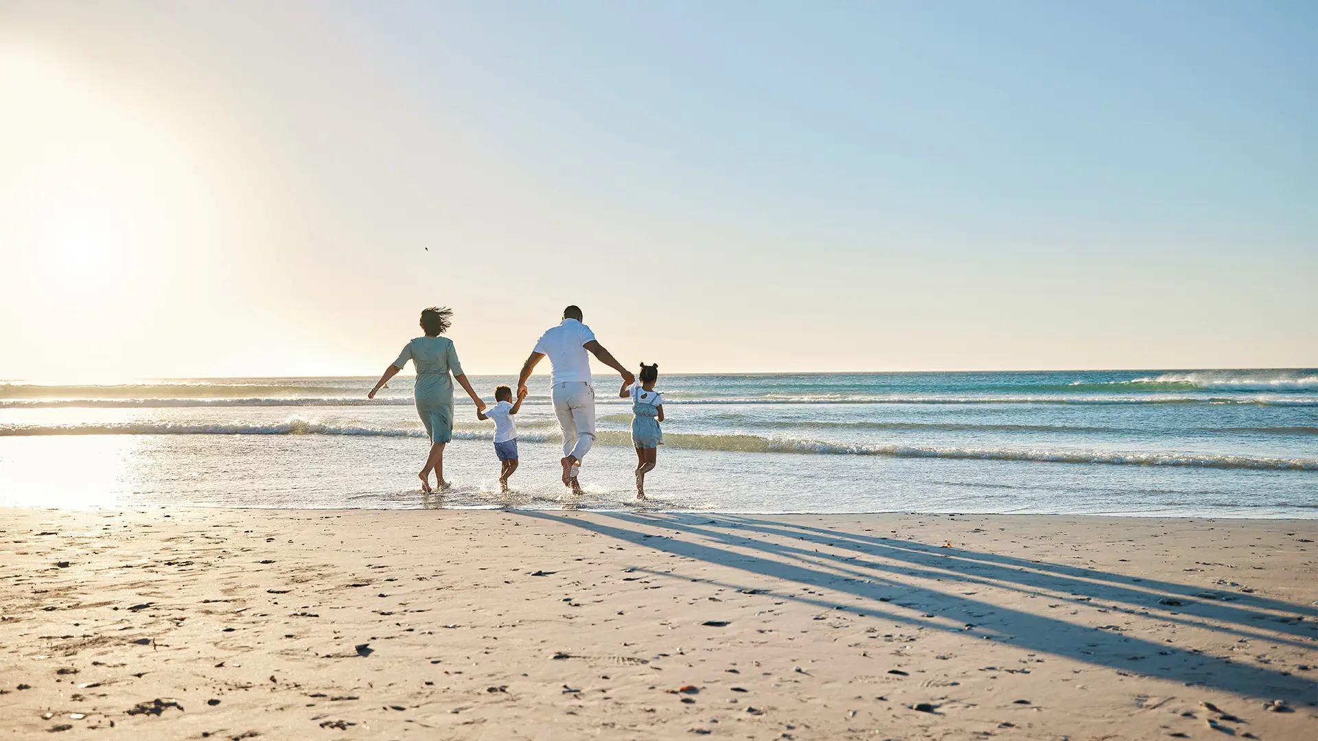 A family of four walks hand in hand along a sunny beach, casting long shadows on the sand as the waves gently lap the shore.