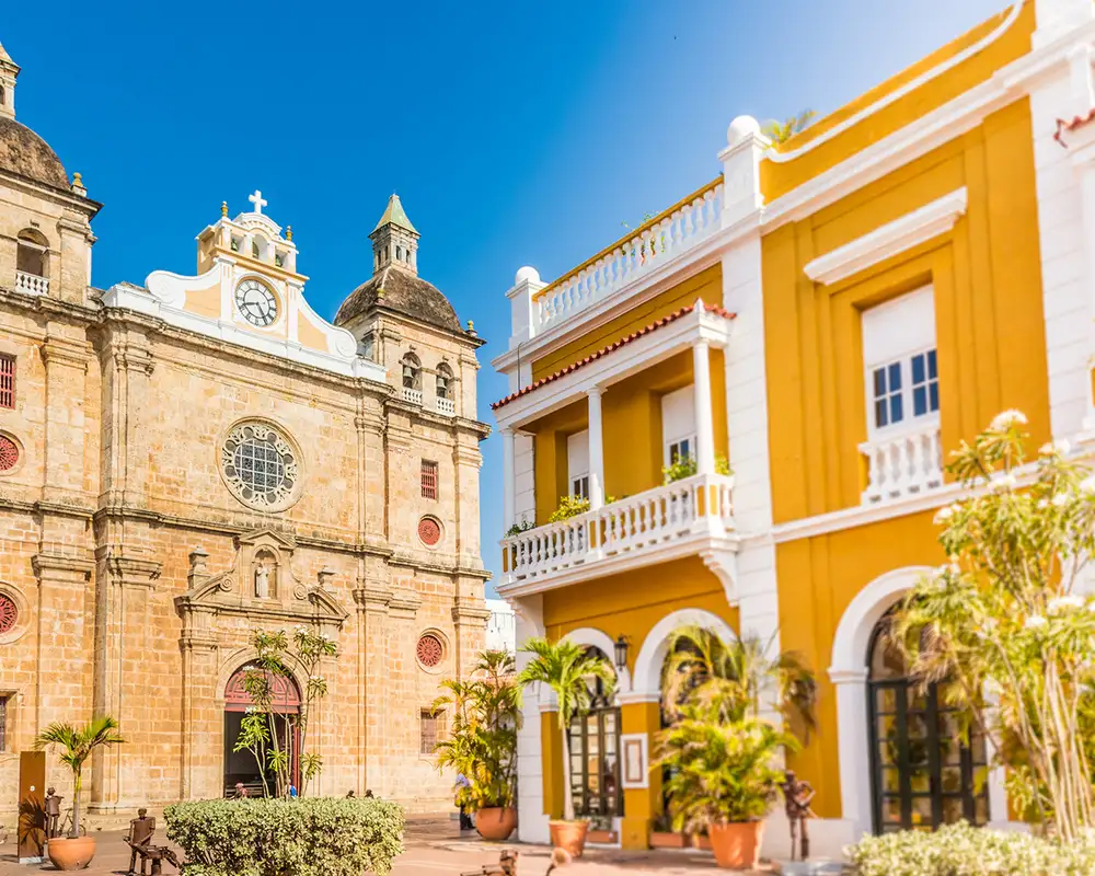 Historic stone church with twin domes beside a vibrant yellow colonial building, set under a clear blue sky.