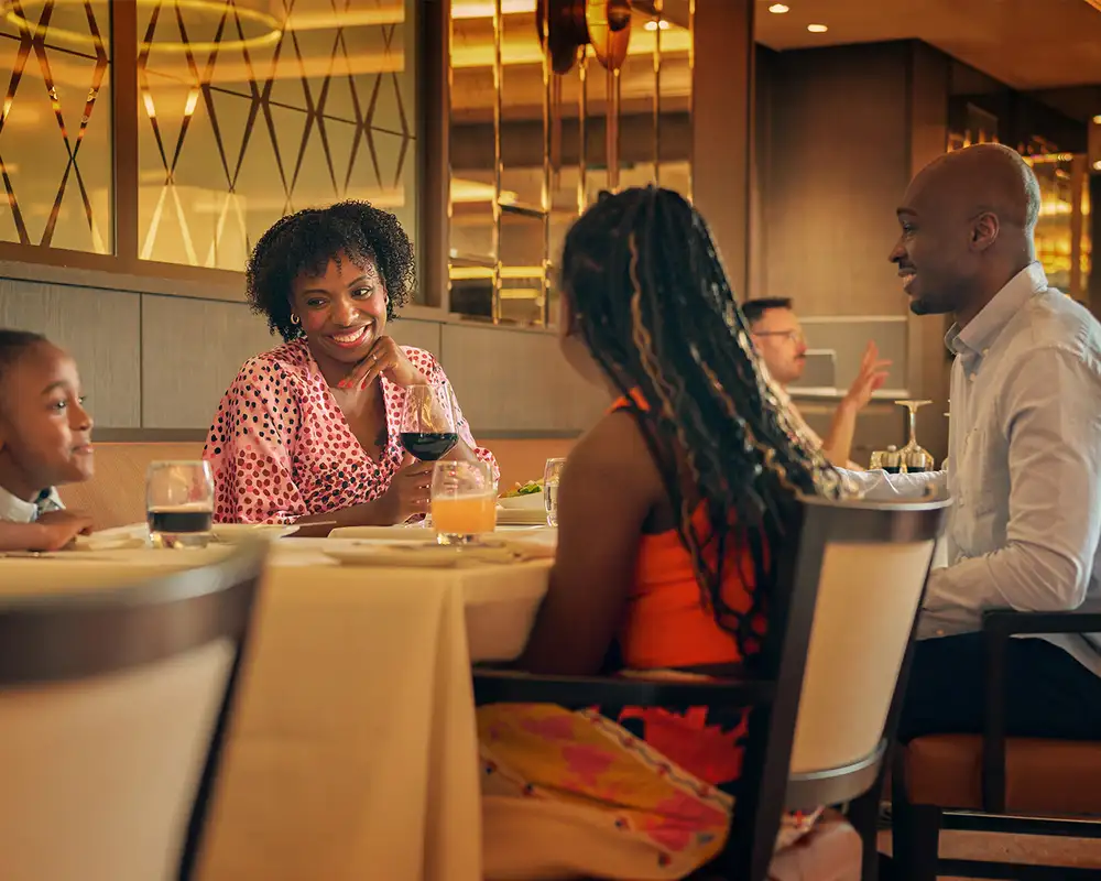 A family of four enjoying a meal together at a restaurant, with a mother smiling and holding a glass of wine, and lively conversation all around. Aboard a P&O Cruise Ship.