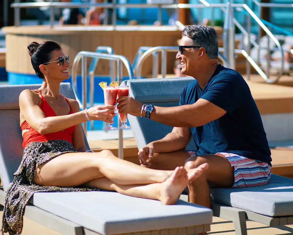 A smiling couple lounging on poolside chairs clink glasses with pink drinks, enjoying a sunny day by the pool. Aboard an Azamara Ship.