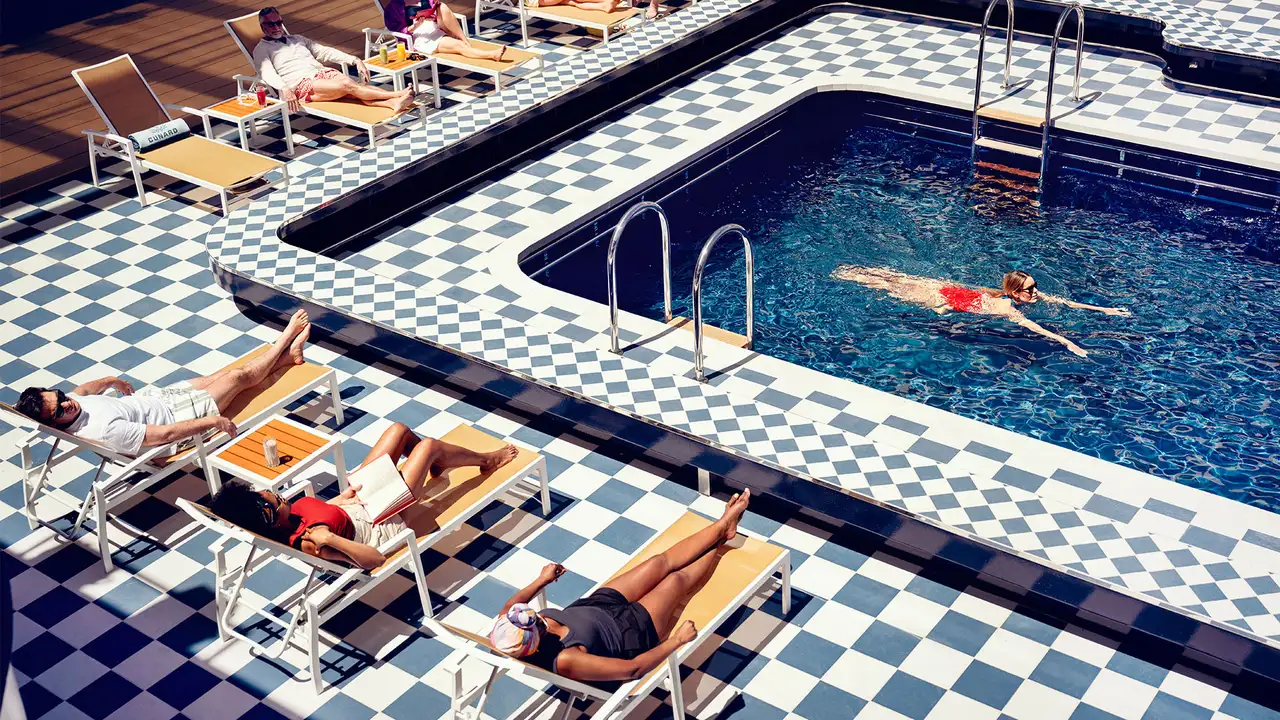 A checker-board patterned pool deck with people in sun chairs and a woman swimming in the pool