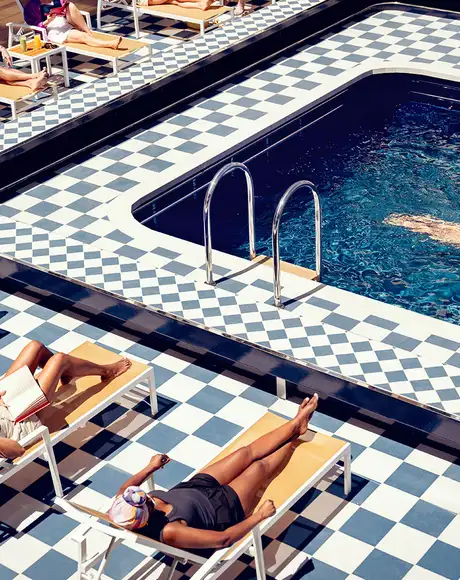 A checker-board patterned pool deck with people in sun chairs and a woman swimming in the pool