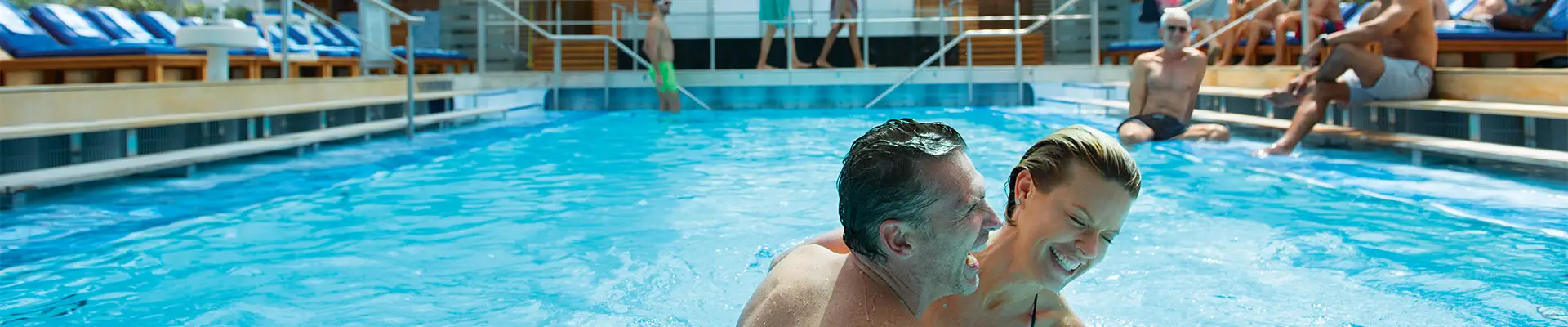 A couple laughing and swimming in an indoor pool, with people relaxing on loungers and walking in the background. Aboard a Celebrity Cruise Ship