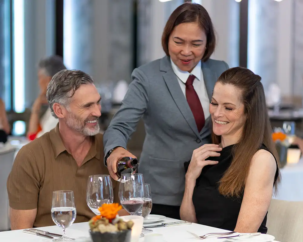 A waiter serving wine to a smiling couple at a restaurant table, with other diners in the background. Aboard an Azamara Cruise Ship.