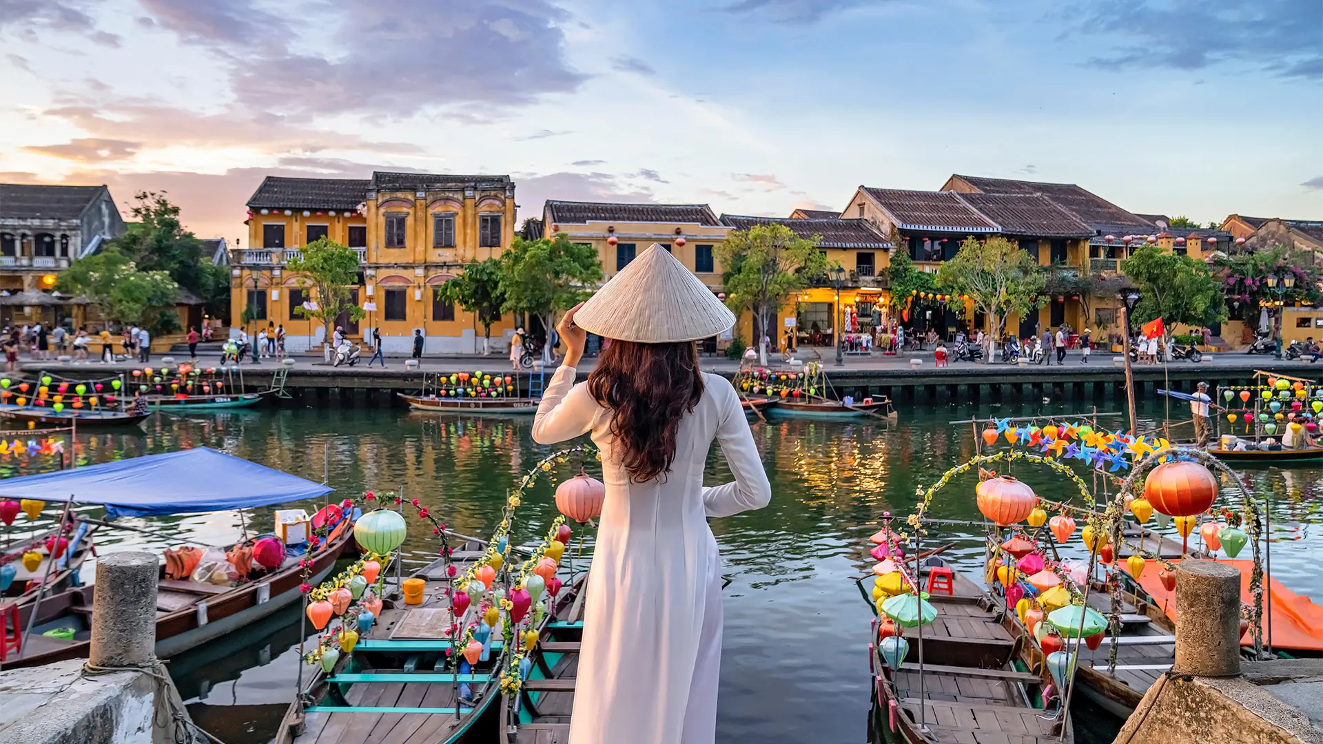 A woman in a white dress and conical hat stands on a bridge, overlooking a canal with colorful lanterns and boats, against a backdrop of old buildings.