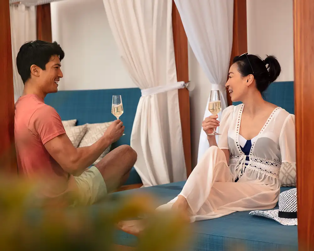A man and woman are sitting on a cushioned daybed under a canopy, toasting with champagne glasses and smiling at each other. Aboard a P&O Cruise Ship.