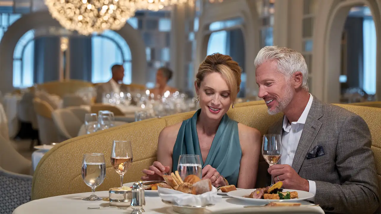 A couple dining elegantly, sharing a meal with wine in a luxurious, well-lit restaurant with a grand chandelier overhead.
