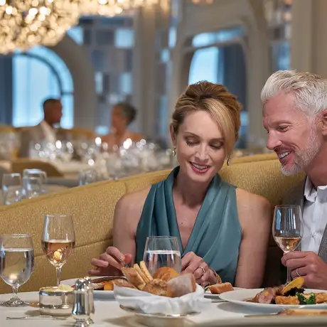 A couple dining elegantly, sharing a meal with wine in a luxurious, well-lit restaurant with a grand chandelier overhead.
