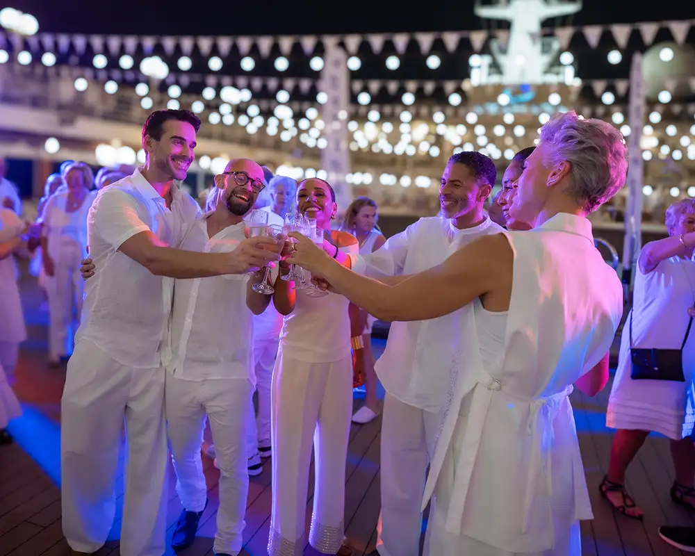 People in white attire celebrating and toasting with glasses on a decorated outdoor deck at night, surrounded by festive lights. Aboard an Azamara Cruise Ship.