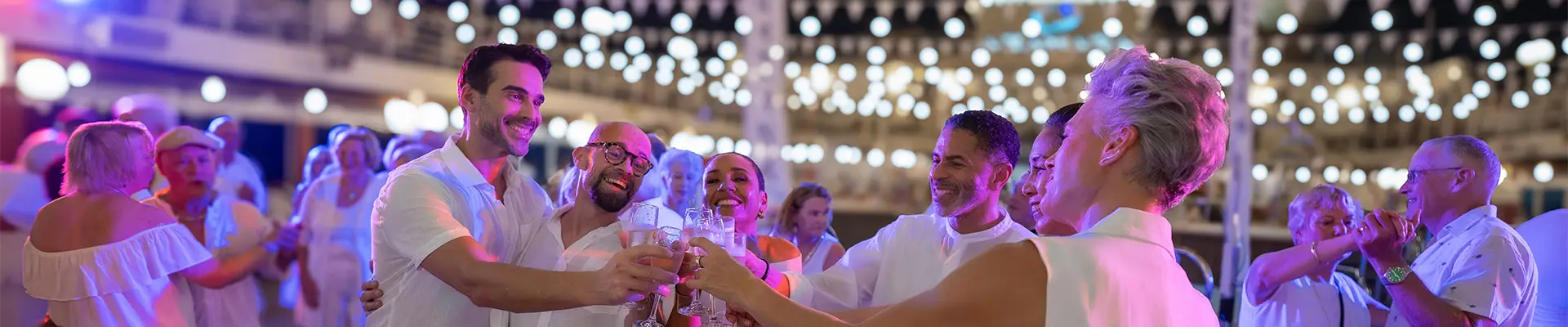 People in white attire celebrating and toasting with glasses on a decorated outdoor deck at night, surrounded by festive lights. Aboard an Azamara Cruise Ship.