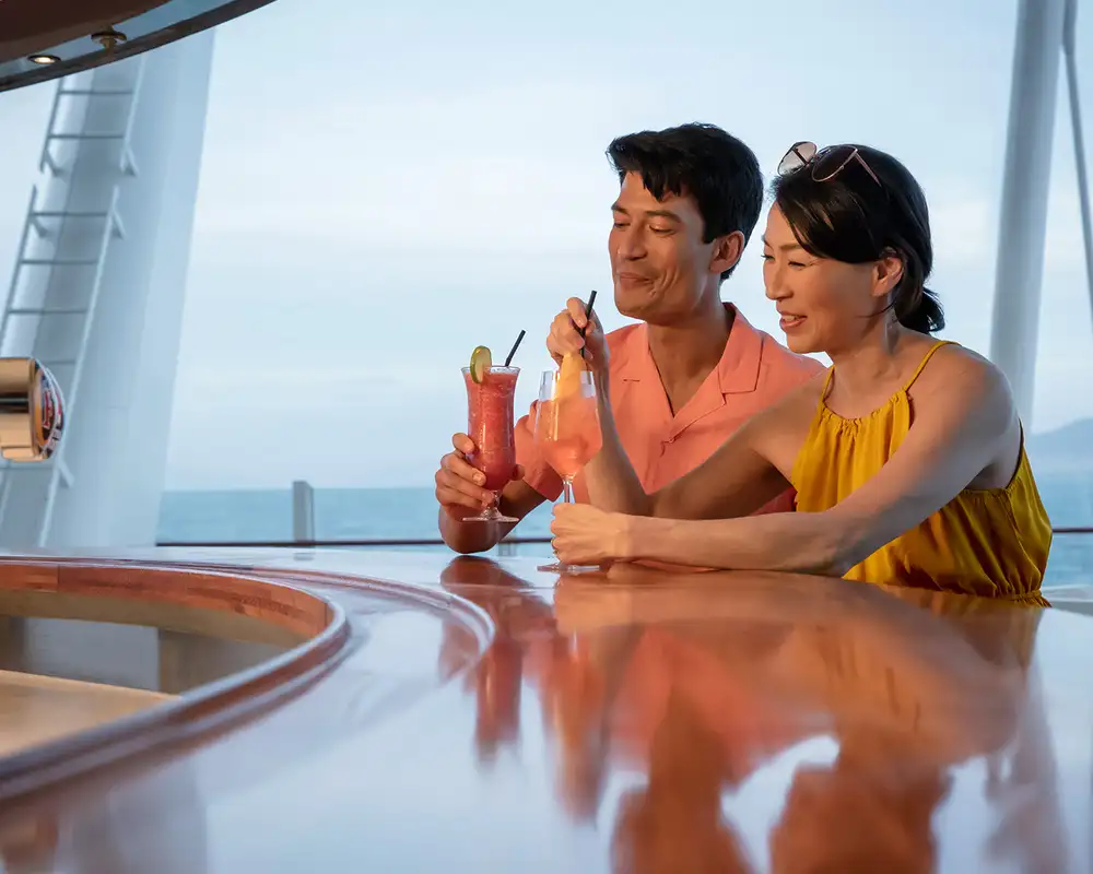 A couple enjoys colorful cocktails at a curved bar on a ship, with the ocean and distant mountains visible through large windows. Aboard a P&O Cruise Ship.