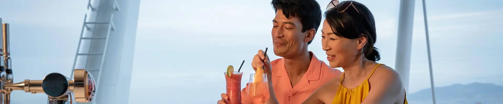 A couple enjoys colorful cocktails at a curved bar on a ship, with the ocean and distant mountains visible through large windows. Aboard a P&O Cruise Ship.