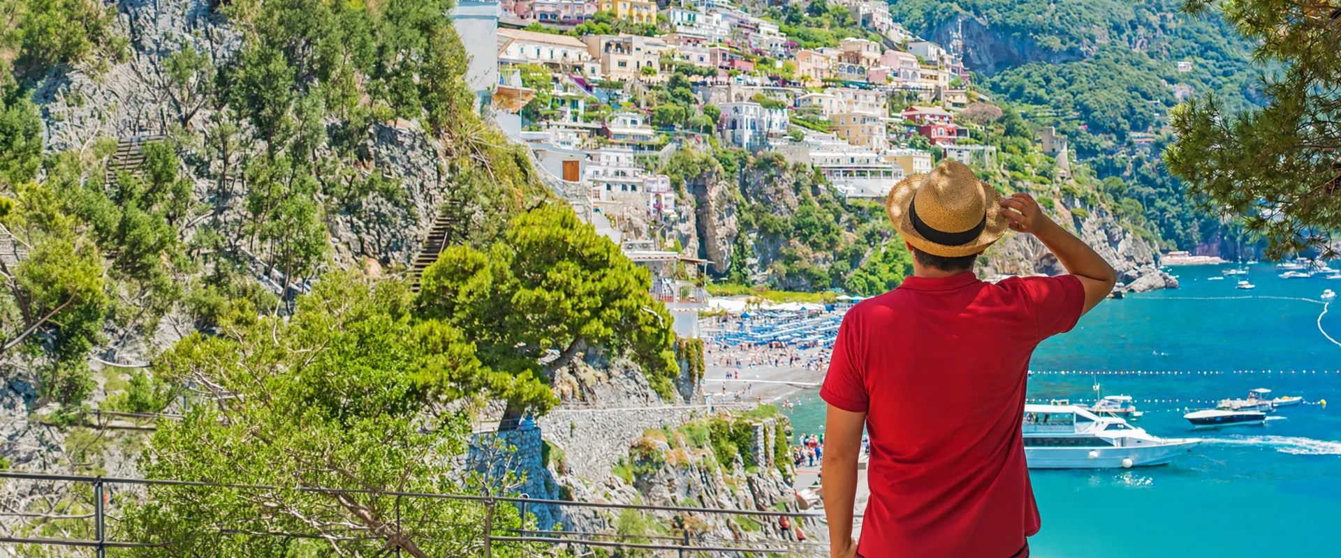 Man in a red shirt and straw hat admires a coastal town with vibrant buildings on a hillside, overlooking a bay with boats and clear blue water.