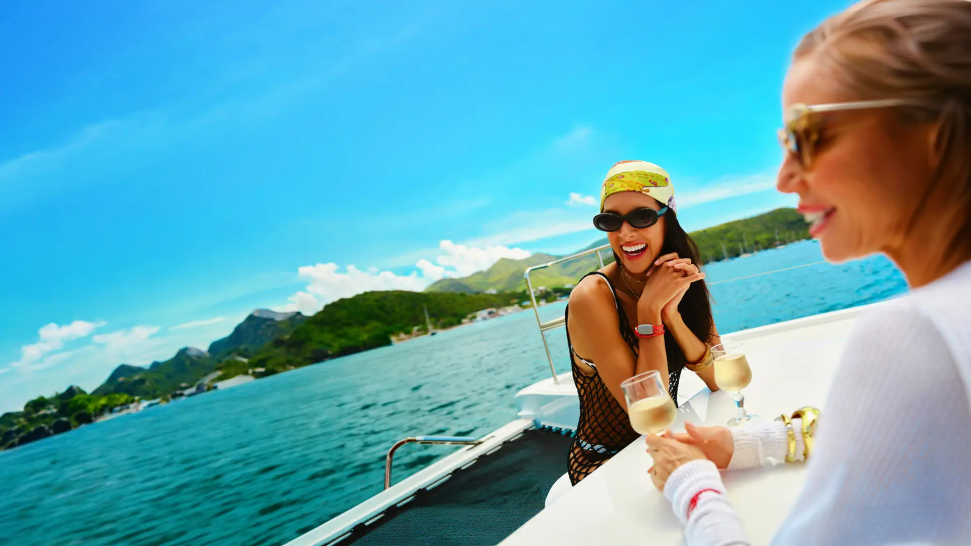 Two women enjoying drinks on a boat with a scenic view of the ocean and green hills under a clear blue sky.