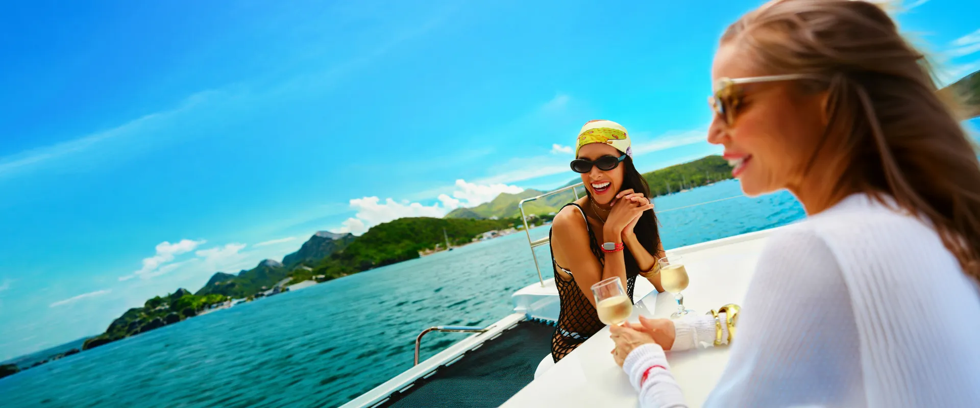 Two women enjoying drinks on a boat with a scenic view of the ocean and green hills under a clear blue sky.
