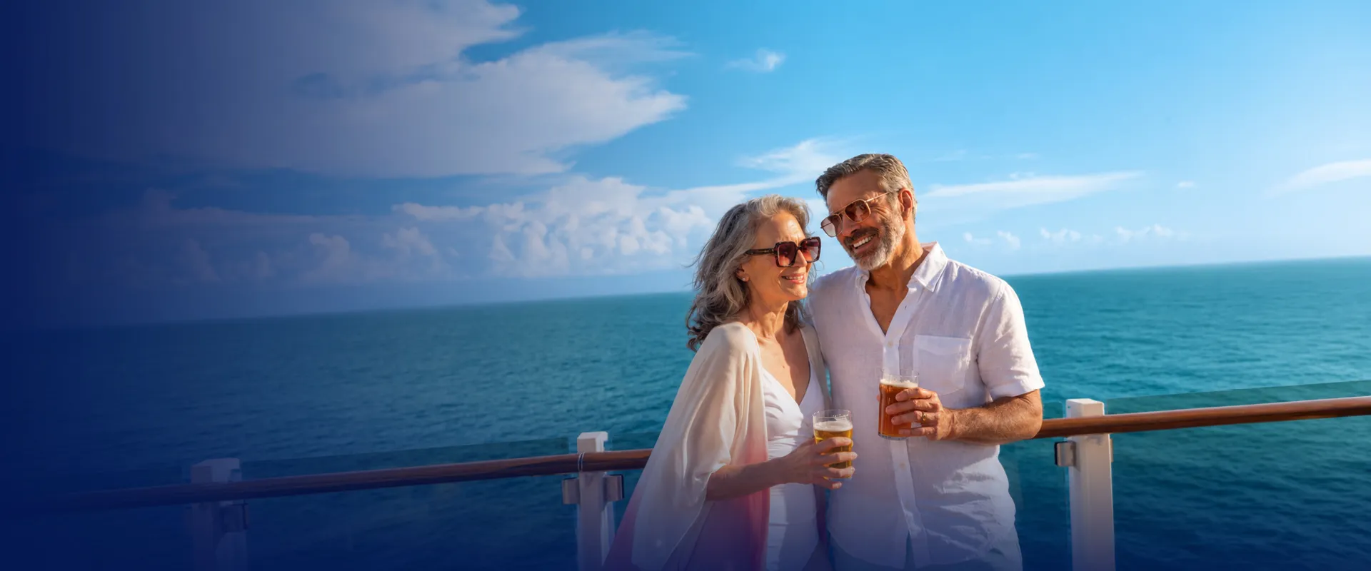 A couple in sunglasses smiles and holds drinks on a cruise ship deck, with the ocean and blue sky in the background.