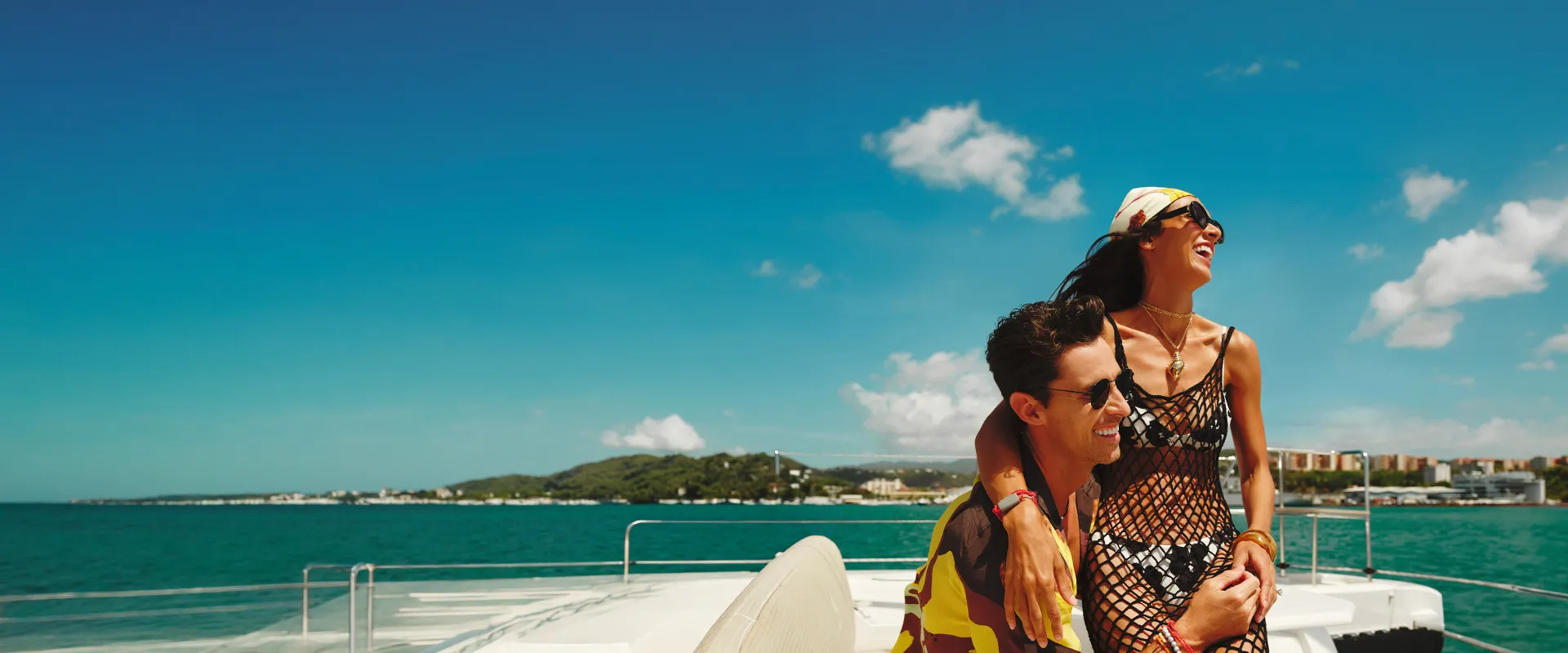 A smiling couple enjoying a sunny day on a boat, with clear blue skies and distant shoreline in the background.