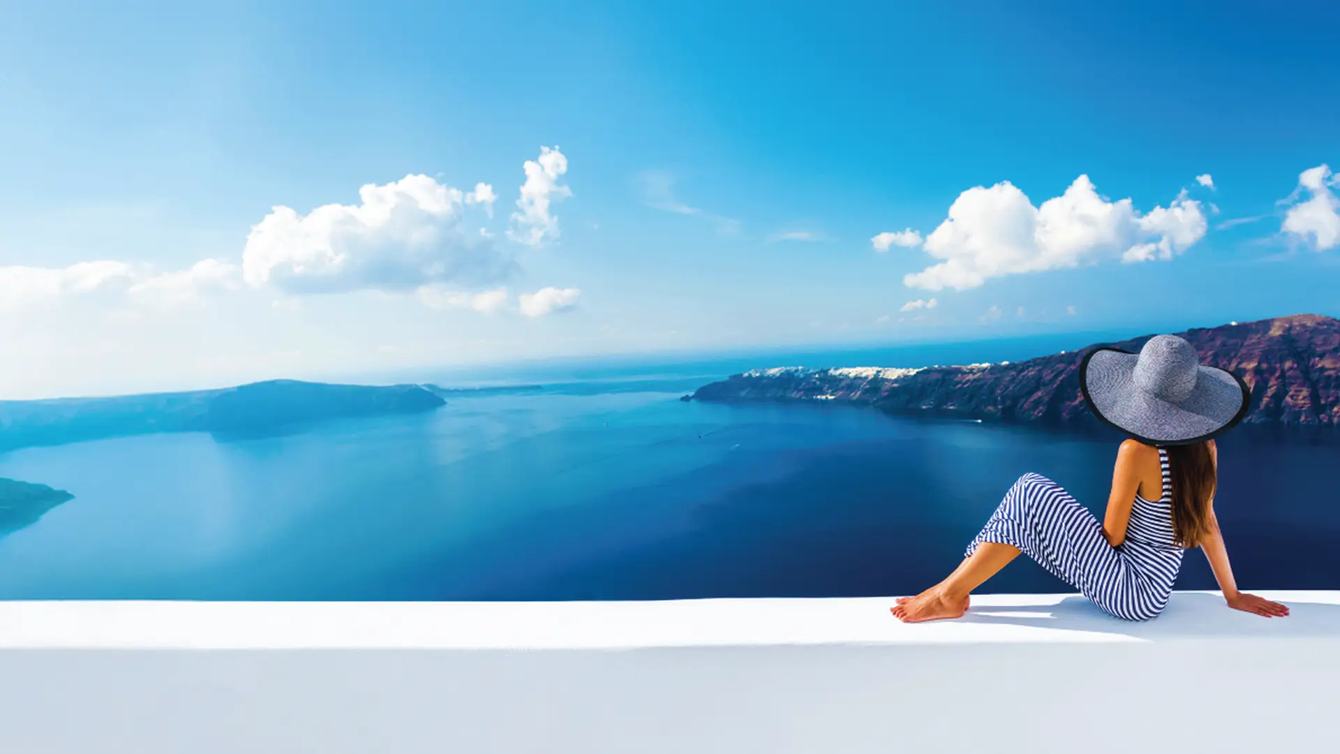 Woman in a wide-brimmed hat sits on a white ledge, overlooking a scenic view of the sea and mountains under a clear blue sky.