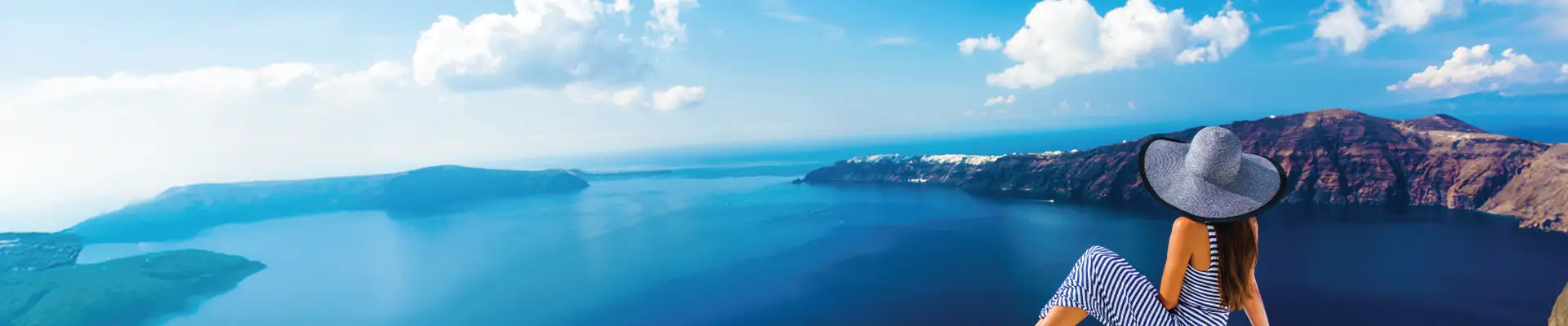 Woman in a wide-brimmed hat sits on a white ledge, overlooking a scenic view of the sea and mountains under a clear blue sky.