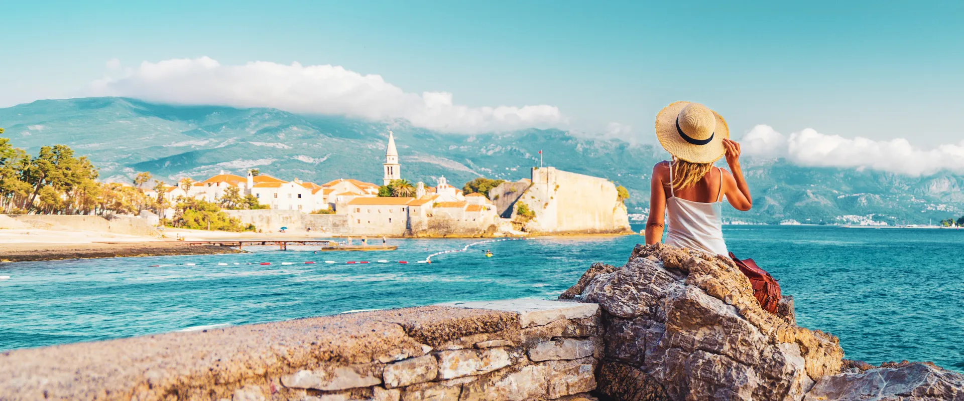 A woman in a sun hat sits on a stone wall, gazing at a coastal town with a church and mountains in the background.