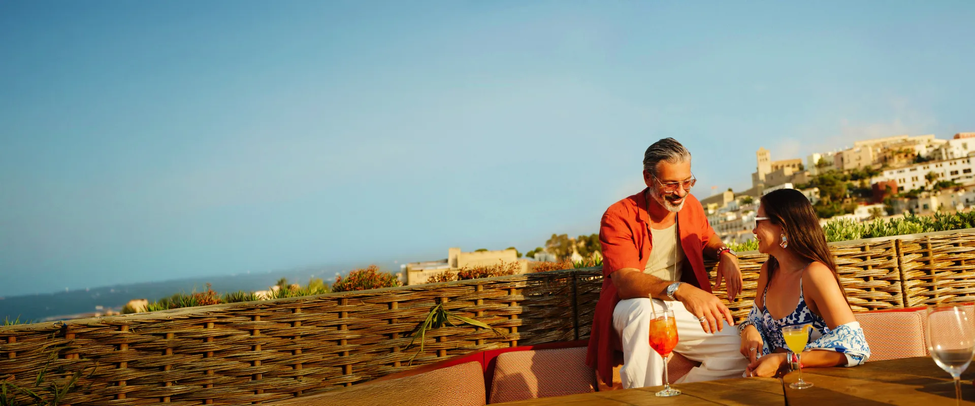 A man and woman enjoy drinks on a sunny terrace overlooking a scenic hillside town, with wicker seating and a clear blue sky.